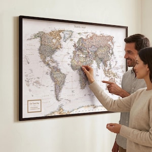 Op de afbeelding: Een wereldkaart met een donkerbruine lijst. De kaart toont landen en geografische kenmerken. Een stel wijst naar de kaart en markeert een locatie. De kaart is bovenaan gemarkeerd met "TRAVEL MAP".