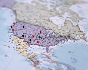 May include: A printed map of North America featuring the United States highlighted in a purple hue. Several small, round push pins in contrasting red and black colors are inserted into various locations across the United States territory.