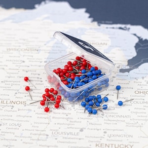 May include: A clear plastic container filled with red and blue push pins, scattered on a map. The pins are small, with round heads and metal shafts. The map shows a detailed view of the Midwestern United States.