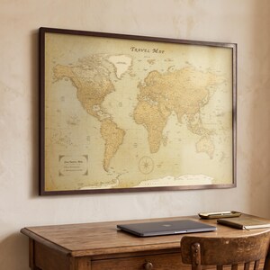 May include: A rectangular wall-mounted world map with a dark brown frame. The map features a vintage-style beige and gold color palette with the text Travel Map at the top and a decorative compass rose in the lower right quadrant of the map.