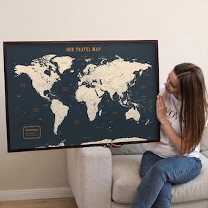 Puede incluir: Un mapa del mundo enmarcado con un fondo azul oscuro y continentes beige. El texto "OUR TRAVEL MAP" está en la parte superior y "The World awaits" en la parte inferior. El mapa está sostenido por una persona.