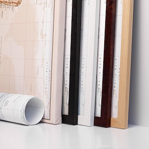 May include: A collection of framed world maps in various wood frame colors, including black, white, and natural wood. A rolled-up map is in the foreground. The maps feature a neutral color palette and are labeled with geographical details.