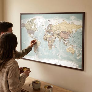 以下が含まれることがあります： 茶色の木製フレームに入った世界地図。 地図は、落ち着いた色と詳細な地理的特徴を備えたヴィンテージの美学を持っています。 2 人が地図を操作し、場所を指しています。 2 つのマグカップの紅茶が木製の表面に置かれています。