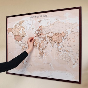 May include: A framed world map with the text "ADVENTURE MAP" at the top. The map is in shades of brown and beige, with red pins marking locations. A person's arm and hand are visible, placing a pin on the map.