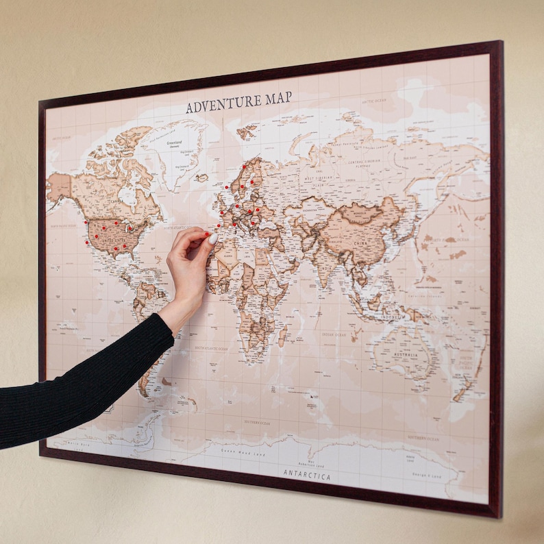 May include: A framed world map with the text "ADVENTURE MAP" at the top. The map is in shades of brown and beige, with red pins marking locations. A person's arm and hand are visible, placing a pin on the map.