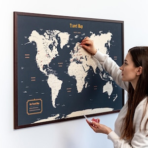 Può includere: Una mappa del mondo di viaggio incorniciata, con sfondo blu scuro e cornice marrone. La mappa presenta masse terrestri beige ed è etichettata con i nomi degli oceani. Una persona sta mettendo un segnaposto sulla mappa. Il testo "Travel Map" è in alto.