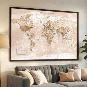 May include: A framed world map with a brown wooden frame. The map is in shades of brown and beige, with the text "Adventure Map" at the top. The map is hanging above a gray sofa with pillows.