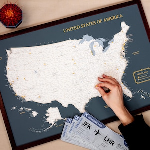 Könnte beinhalten: Eine gerahmte Karte der Vereinigten Staaten von Amerika in Marineblau mit goldfarbenen Buchstaben und Pins zur Markierung von Orten. Die Karte enthält den Text "UNITED STATES OF AMERICA" und "TRAVEL MAP". Drei Bordkarten sind sichtbar, mit Flugdetails von JFK nach LHR.