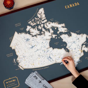 May include: A dark teal world map of Canada with a wooden relief. The map is labeled with the names of oceans and geographical features. A person is placing a pin on the map. A boarding pass is visible.