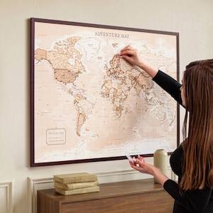 Può includere: Una mappa del mondo incorniciata, intitolata "ADVENTURE MAP", nei toni del marrone e beige. Spilli rossi segnano le località sulla mappa. La cornice marrone scuro circonda la mappa. La mappa è esposta su un muro.