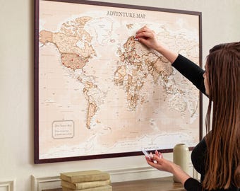 Op de afbeelding: Een ingelijste wereldkaart getiteld Adventure Map met een lichtbeige achtergrond en bruine continenten. De kaart bevat rode spelden die verschillende locaties over de hele wereld markeren, met een klein vakje linksonder met Our Travel Map.