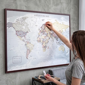 May include: A framed world map with the text "ADVENTURE MAP" at the top. The map is in neutral tones with red pins marking locations. A person is placing a pin on the map. The frame is a dark brown color.
