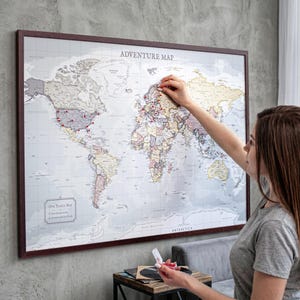 May include: A framed world map with the text "ADVENTURE MAP" at the top. The map is light-coloured with country outlines and red pins marking locations. A person is placing a pin on the map.