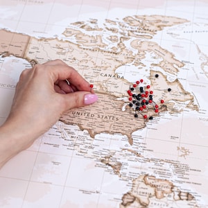 May include: A world map with a vintage aesthetic, featuring the United States and Canada. Red and black push pins mark locations. A hand with pink nail polish places a red pin on the map, highlighting a specific spot.
