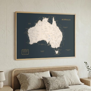 May include: A framed map of Australia, featuring a dark blue background with a detailed beige map. The word "AUSTRALIA" is printed in gold at the top. The map is in a light wood frame, hanging above a bed with pillows.