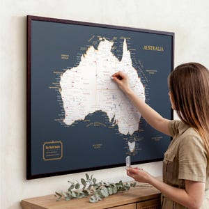 May include: A framed map of Australia with a dark blue background and white landmass. The map is labelled with gold text and includes push pins. The frame is dark brown. The text "The World Awaits" is in the lower left corner.