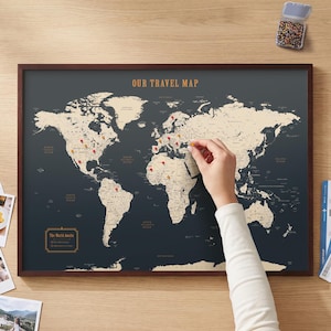 Pode incluir: Um mapa-múndi emoldurado com fundo cinza-escuro e continentes na cor creme. O texto "OUR TRAVEL MAP" está impresso no topo e uma caixa com o rótulo "The World Awaits" está no canto. Tachinhas vermelhas e amarelas marcam locais no mapa.