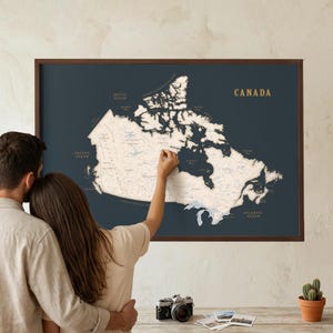 May include: A framed map of Canada with a dark blue background and a detailed white outline of the country. The word "CANADA" is printed in gold. A couple is looking at the map, with one person pointing at it.