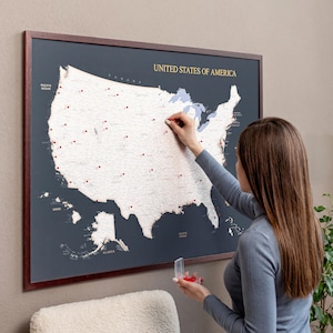 May include: A framed wall map of the United States of America with a dark gray background and a light-colored map surface. The map features red push pins placed across various states, and the text UNITED STATES OF AMERICA is printed in gold.