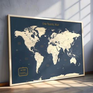 May include: A framed world map with a navy blue background and gold-colored continents. The map is labeled "OUR TRAVEL MAP" in gold lettering. A small gold-framed section reads "Jake's Travels". The map is displayed on a wooden floor.