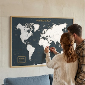 May include: A framed world map with a dark blue background and white continents. The map is labelled "OUR TRAVEL MAP" in gold lettering. A couple is shown placing pins on the map, indicating travel destinations. The frame is made of light wood.