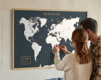 May include: A framed world map with a dark blue background and white continents. The map is labeled "OUR TRAVEL MAP" in gold lettering. A couple is shown placing pins on the map, indicating travel destinations. The frame is made of light wood.