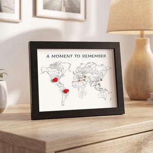 May include: A black framed wall art piece featuring a white world map with the text A MOMENT TO REMEMBER printed at the top. The map includes a red house-shaped pin, a red heart-shaped pin, and several small metallic push pins across the continents.