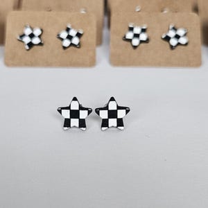 May include: A pair of star-shaped stud earrings with a black and white checkered pattern. The earrings are displayed on a white surface, with several other pairs visible in the background, mounted on small brown cards.
