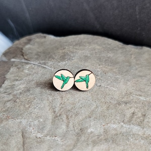 May include: A pair of wooden earrings with a green hummingbird design on a light brown background.