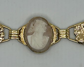 May include: A gold bracelet with a cameo and floral accents. The cameo features a woman's profile in white against a light brown background. The flowers are gold and have a delicate design.
