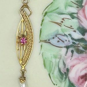 May include: A gold pendant featuring an elongated, openwork marquise shape with a central pink gemstone. The pendant includes a textured, curved side element and a small, dangling white pearl at the bottom, suspended from a gold bail.