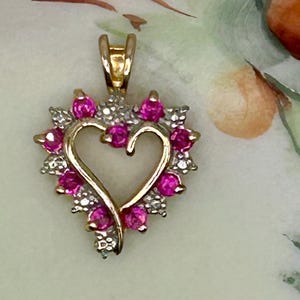 May include: A gold-tone heart pendant with a halo of pink gemstones and small white stones. The pendant features a heart-shaped design with a bail for a chain. The gemstones are set around the heart, creating a sparkling effect.