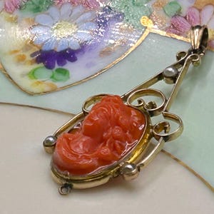 May include: A gold pendant featuring a carved orange cameo of a woman's profile. The pendant has a decorative gold frame with scrollwork and small pearl accents. The background is a light green and white floral design.