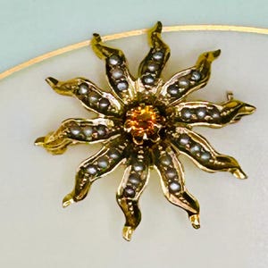 May include: A gold-toned floral brooch with a central orange gemstone and petal-like structures adorned with small, white pearls. The brooch is set against a soft, off-white background, creating a striking contrast.