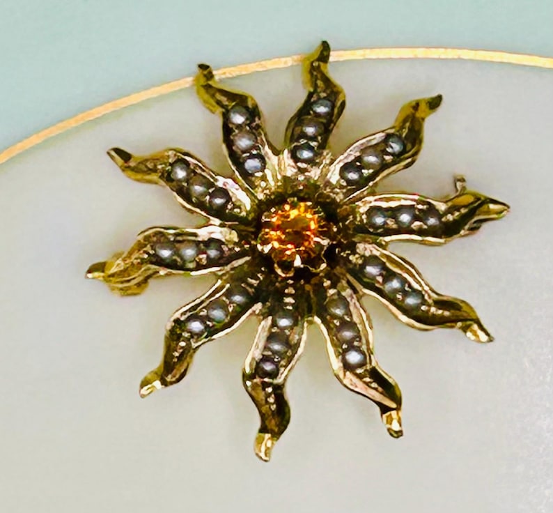 May include: A gold-toned floral brooch with a central orange gemstone and petal-like structures adorned with small, white pearls. The brooch is set against a soft, off-white background, creating a striking contrast.