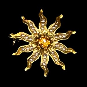 May include: A gold-toned sunburst brooch with a central orange gemstone and surrounding white pearls. The brooch has a pin clasp on the back and is set against a black background. The sunburst design features wavy rays.