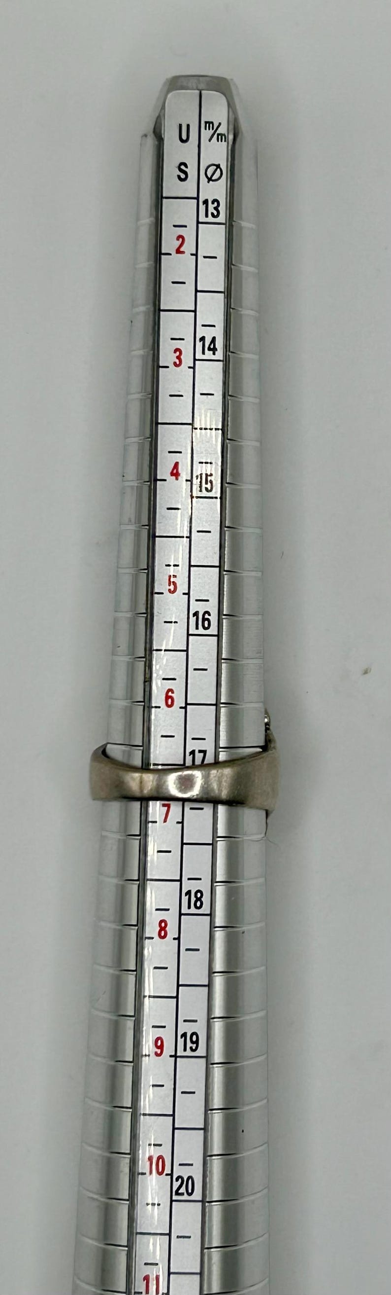 May include: A silver ring size gauge with markings from size 13 to 20. The gauge is used to measure the size of a ring.