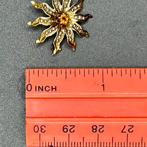May include: A gold-toned flower-shaped brooch with a central orange gemstone and small white pearls around the petals. The brooch is approximately 1 inch in diameter. The brooch is on a gray surface next to an orange ruler.
