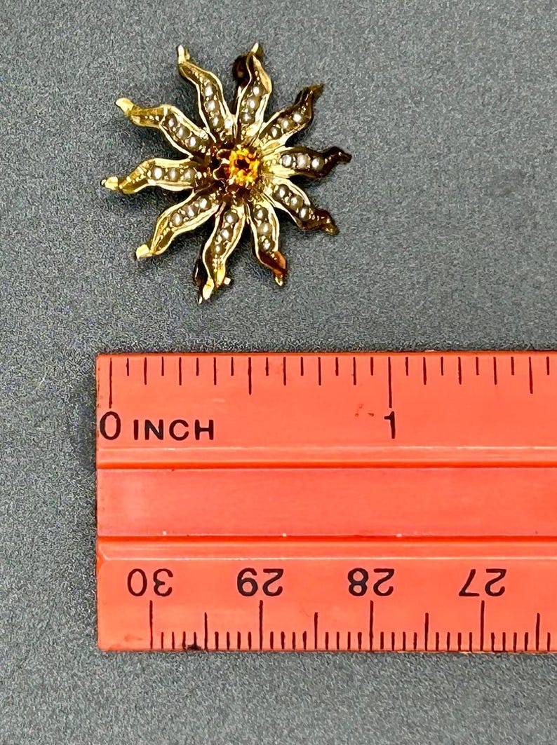 May include: A gold-toned flower-shaped brooch with a central orange gemstone and small white pearls around the petals. The brooch is approximately 1 inch in diameter. The brooch is on a gray surface next to an orange ruler.