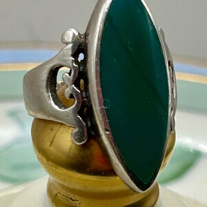 May include: A silver ring with an ornate design and a large, dark green oval stone.