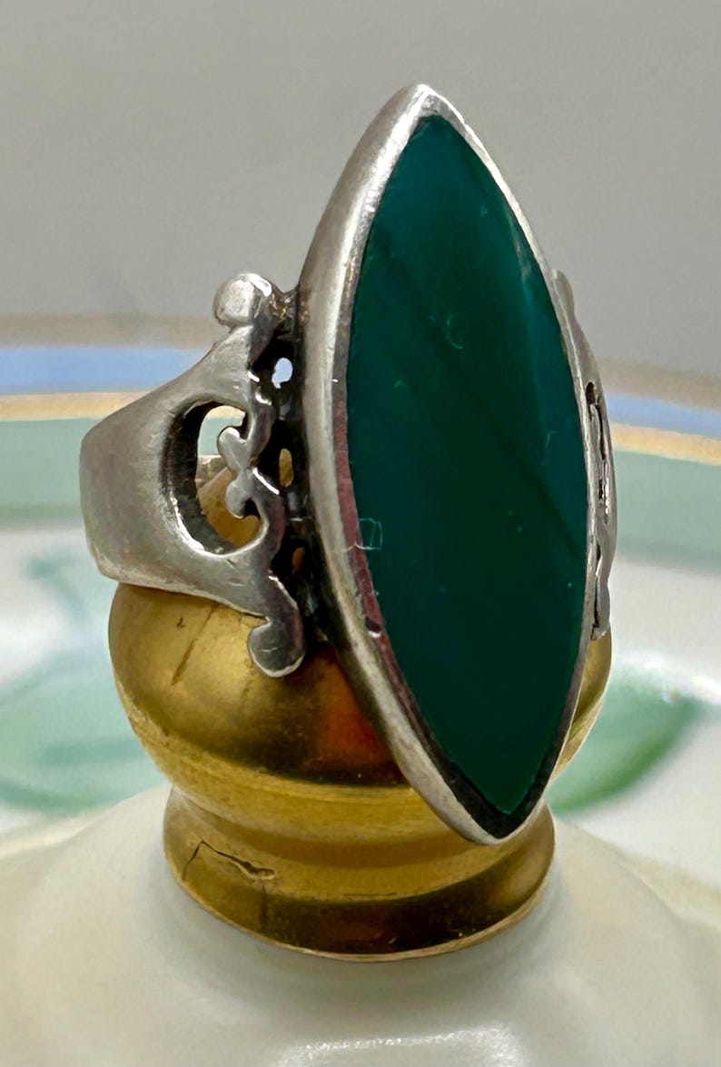 May include: A silver ring with an ornate design and a large, dark green oval stone.