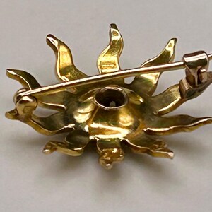 May include: A gold-toned sunburst brooch with a pin closure. The brooch features a detailed design with radiating rays and a central setting. The back of the brooch shows the pin mechanism and a small, dark-colored stone.