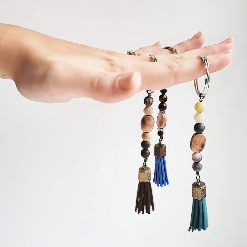 May include: Three beaded keychains with tassels. The keychains feature different colored beads and tassels, including brown, black, white, blue, and green.