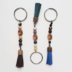 May include: Three keychains with different colored tassels and beaded charms. The charms are made of natural stones and metal. The tassels are brown, blue, and teal.