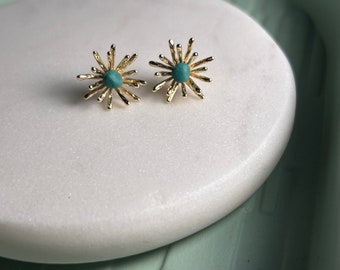 May include: A pair of gold-coloured stud earrings. Each earring features a turquoise-coloured centre stone surrounded by a starburst design. The earrings are displayed on a white, oval-shaped surface.