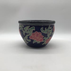 May include: A round ceramic bowl with a deep blue base colour, featuring a repeating pattern of pink flowers and light green leafy vines. The bowl has a smooth, dark, solid-coloured rim at the top and a curved, tapered body shape.