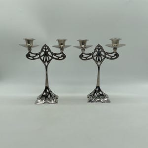 May include: A pair of silver-toned metal candelabras, each featuring two candle holders supported by a central stem with an intricate, openwork, flowing design. The bases are wide and flared, mirroring the decorative pattern of the upper arms.