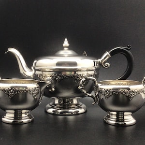 May include: A silver tea set comprising a teapot, creamer jug, and sugar bowl. The teapot features a black handle and a decorative finial. Each piece is embellished with a grape vine pattern around the edges. The set is presented on a dark background.