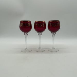 May include: Three wine glasses with deep red bowls featuring a gold-coloured etched grape and leaf pattern around the rim. Each glass has a clear, twisted glass stem and a circular base, standing upright in a row against a plain white background.