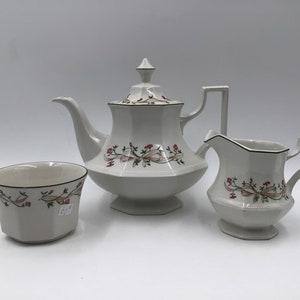 May include: A white porcelain tea set comprising a teapot, creamer jug, and sugar bowl. The set is decorated with pink and tan floral and ribbon motifs, and black trim. The teapot has a lid and handle.